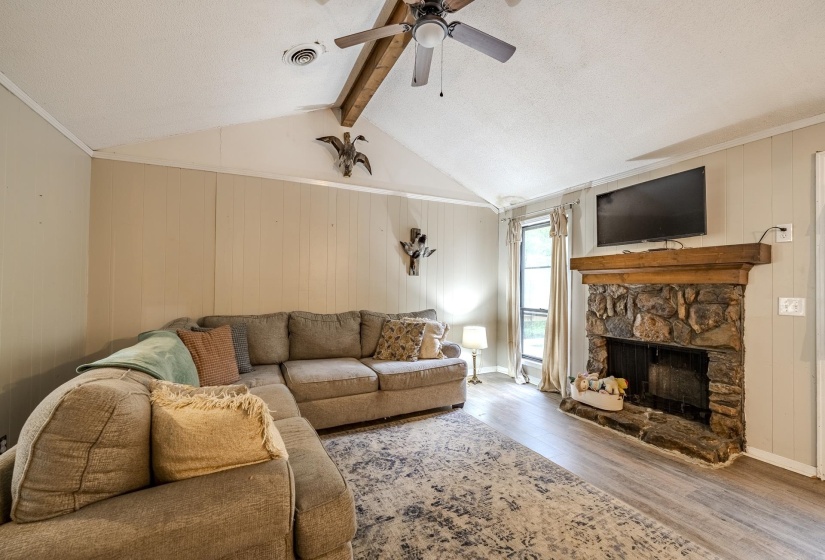 Living area featuring a stone-clad fireplace with a wood mantel, vaulted ceiling with exposed wood beam, and a ceiling fan