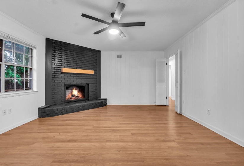 Open-concept living area featuring a prominent dark brick fireplace with a light-toned wood mantel, wood-finish flooring, a contemporary ceiling fan with integrated lighting, white paneling, and an expansive window