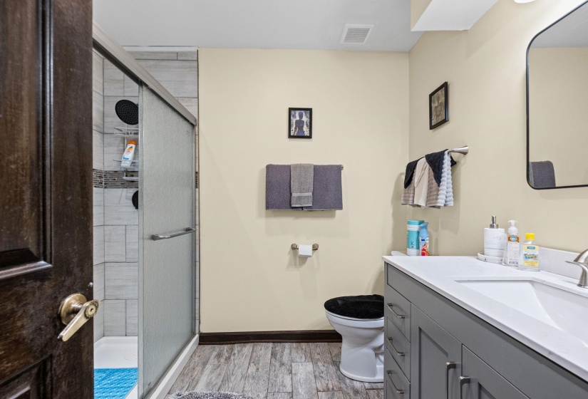 Bathroom featuring a gray vanity with a white countertop and integrated sink, a dark framed mirror, wood-finish flooring, and a tiled shower with a sliding glass door