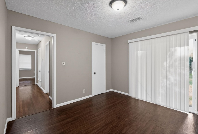 Spacious room featuring wood-finish flooring, neutral-toned walls, a ceiling-mounted light fixture, and vertical blinds covering a glass sliding door