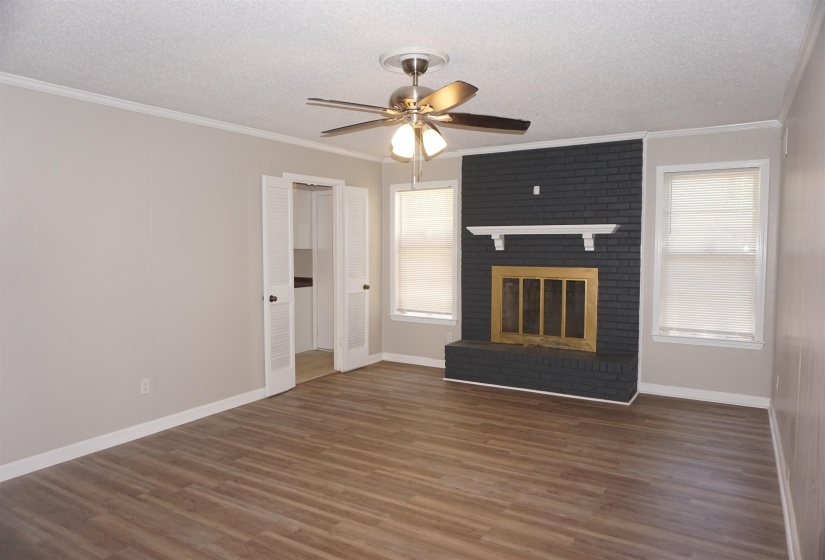 Spacious room featuring wood-finish flooring, a ceiling fan with integrated lighting, and a painted brick fireplace with a white mantel and brass-toned firebox insert