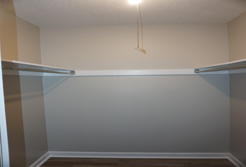 Interior closet featuring wood-finish flooring, light grey and beige painted walls, white baseboards, and built-in white shelving with metal hanging rods