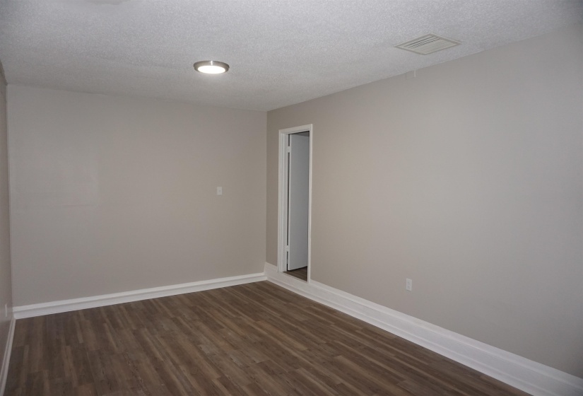 Spacious room featuring wood-finish flooring, light neutral wall paint, white baseboards, a flush-mount ceiling light fixture, and a white ceiling vent