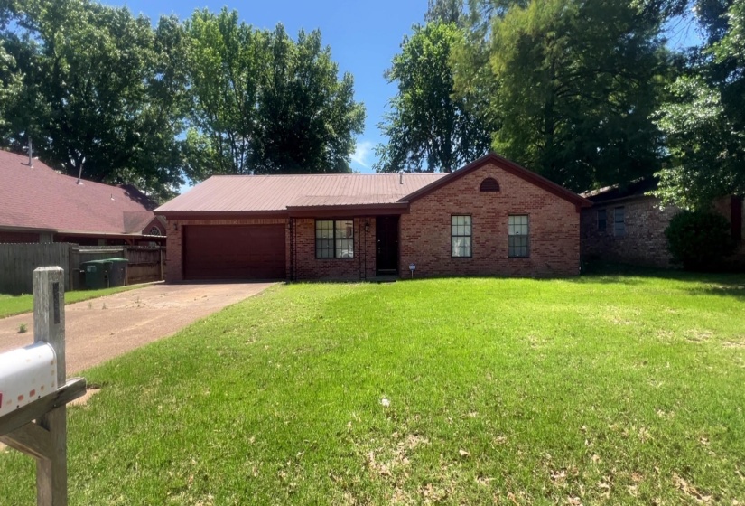 Brick exterior with a red metal roof, featuring a paved driveway, front lawn, and mature trees