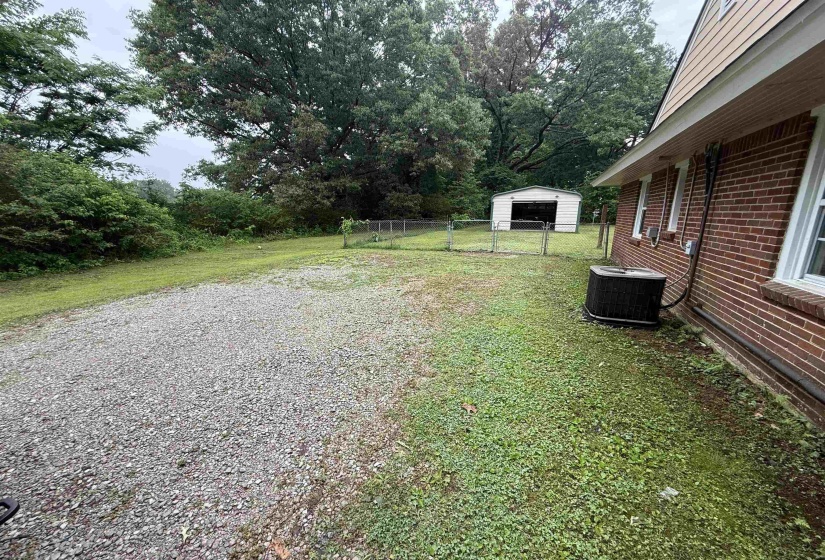 Rear exterior featuring a brick facade, gravel driveway, chain-link fencing, a detached storage shed, and mature perimeter trees
