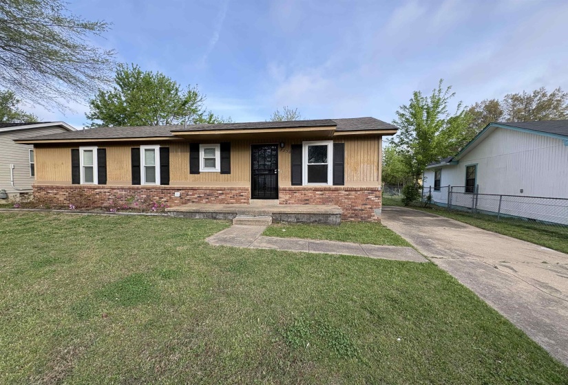 Single-story residence featuring a brick and wood-panel facade, black shutters, a concrete driveway, and a front yard with mature trees