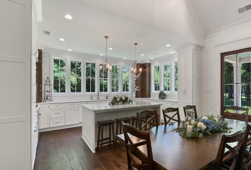 Open-concept kitchen and dining area featuring dark wood-finish flooring, white cabinetry, a central island with countertop seating, and multiple large windows