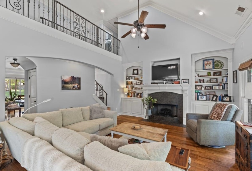 Two-story living area featuring vaulted ceilings, hardwood flooring, a gas fireplace with a stone surround, and built-in shelving