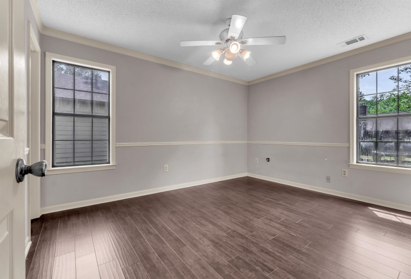 Spacious room featuring wood-finish flooring, two large windows, a ceiling fan with light fixture, crown molding, and chair rail detailing