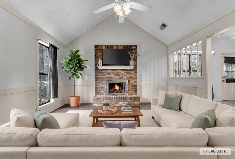 Vaulted ceiling living area featuring a brick fireplace with mantel, dark wood-finish flooring, white wainscoting, and a ceiling fan