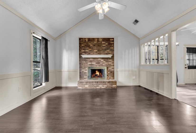 Vaulted ceilings enhance this open-concept room featuring wood-finish flooring, wainscoting, and a brick fireplace with a mantle