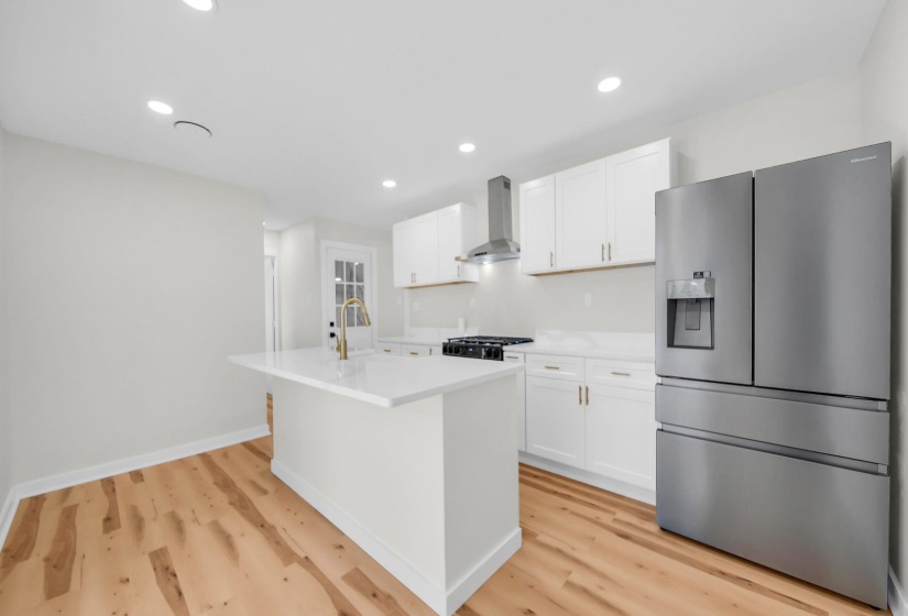 Kitchen featuring stainless steel fridge, a center island with sink, white cabinets, wall chimney range hood, and recessed lighting