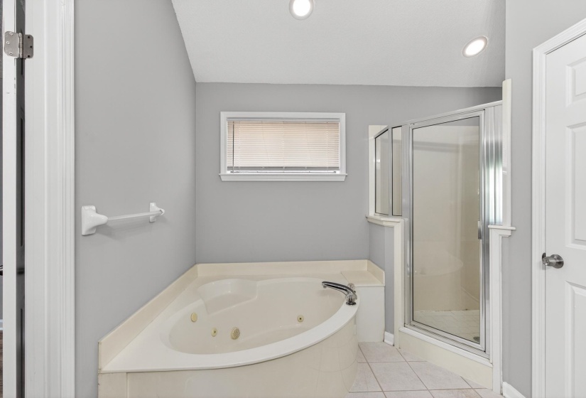 Corner jetted tub with tile surround, glass-enclosed shower, transom window with blinds, and recessed ceiling lighting