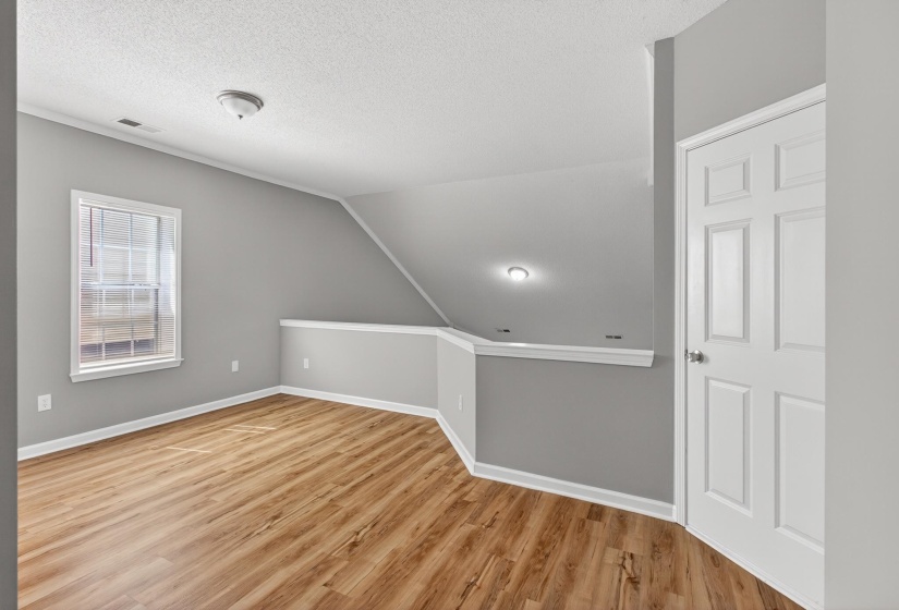 Open loft area featuring wood-finish flooring, light gray wall paint, a white six-panel door, and a white-trimmed window