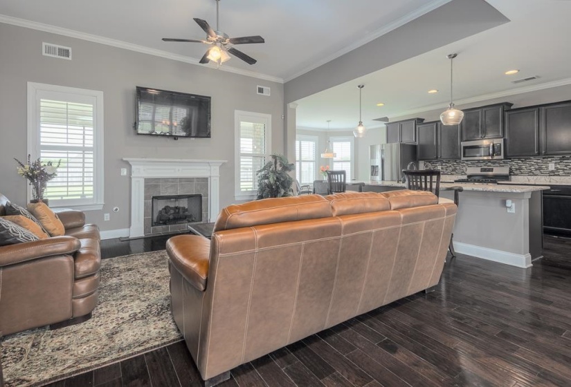Open-concept living area featuring dark wood-finish flooring, a ceiling fan with integrated lighting, and a fireplace with a white mantel and tile surround