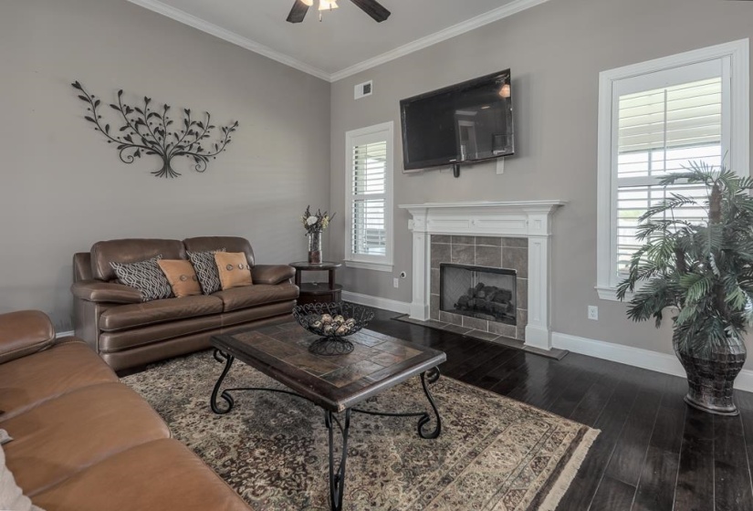 Living area featuring a fireplace with a white mantel and tile surround, dark wood-finish flooring, white window shutters, crown molding, and a ceiling fan