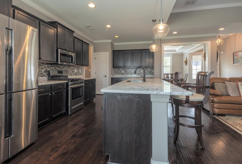 Spacious kitchen featuring an extended island with stone countertop, dark wood-finish flooring, stainless steel appliances, dark cabinetry, and a mosaic tile backsplash