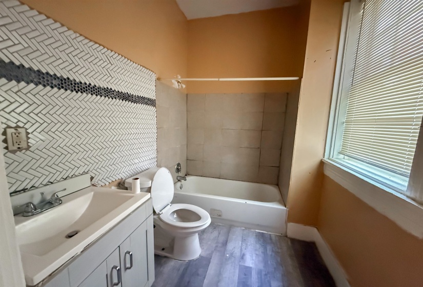 Bathroom featuring a white subway tile backsplash in a herringbone pattern with a dark accent stripe, a single vanity with a white basin and grey cabinetry, a white toilet, a white bathtub with light-colored wall tiling, and wood-finish flooring