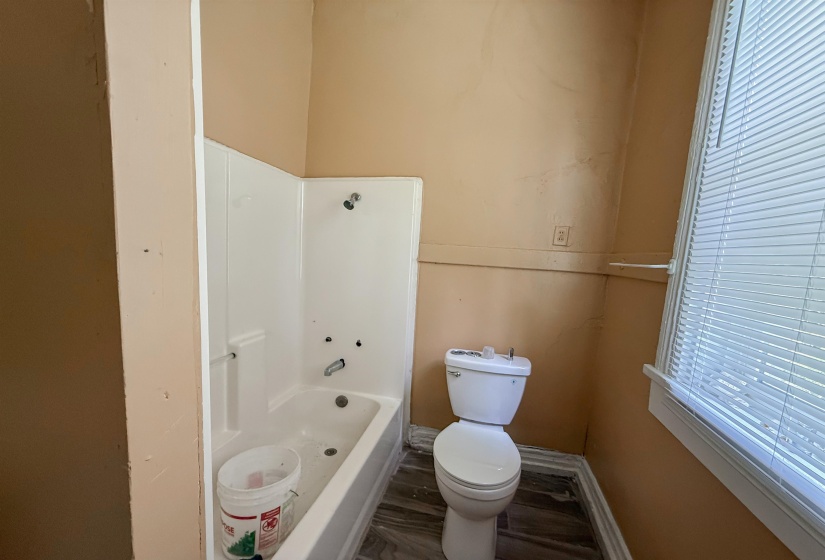 Bathroom featuring a white tub/shower combination, standard toilet, wood-finish flooring, and a window with blinds