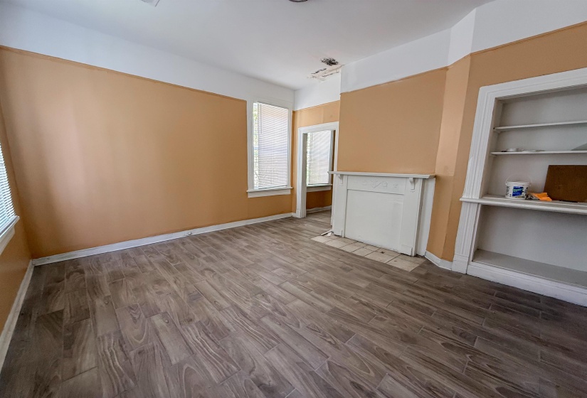 Spacious room featuring wood-finish flooring, two large windows with blinds, a decorative mantelpiece, and built-in shelving