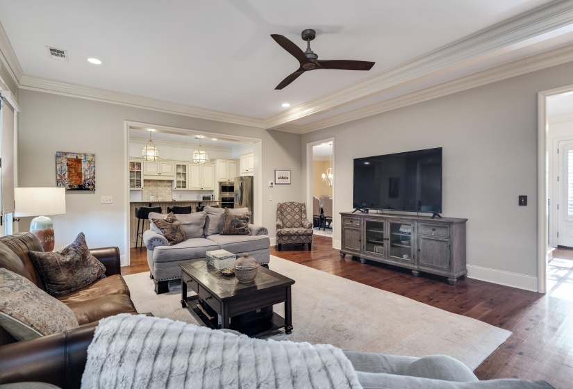 Open concept living area featuring hardwood flooring, crown molding, a ceiling fan, recessed lighting, and a sightline into a kitchen with pendant lighting