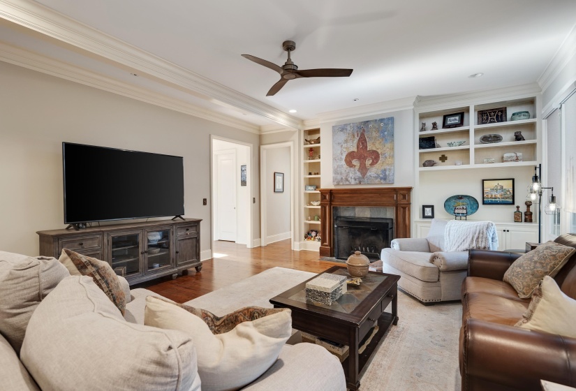 Living area featuring hardwood flooring, a fireplace with a wood mantel, built-in shelving, crown molding, and a ceiling fan