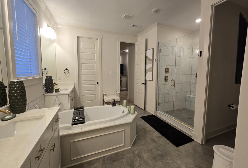 Spacious bathroom featuring a built-in soaking tub, dual vanities with light countertops, a glass-enclosed shower with tile surround and built-in bench, and light gray tile flooring