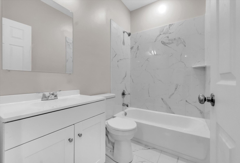 Bathroom featuring a white vanity with integrated sink, chrome faucet, wall-mounted mirror, and a combination shower/tub with marble-look tile surround and a built-in shelf