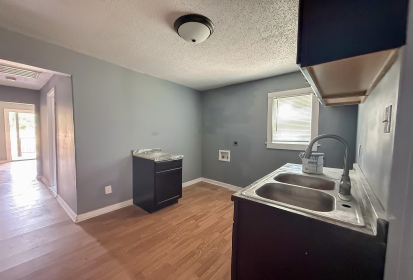 Utility area featuring wood-finish flooring, a double basin stainless steel sink with a gooseneck faucet, a dark base cabinet with a light-toned countertop, and connections for laundry appliances