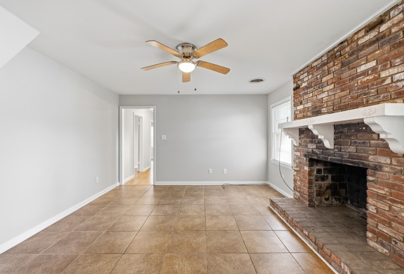 Spacious living area featuring a brick-faced fireplace with an ornate white mantel, ceramic tile flooring, and a ceiling fan with integrated lighting