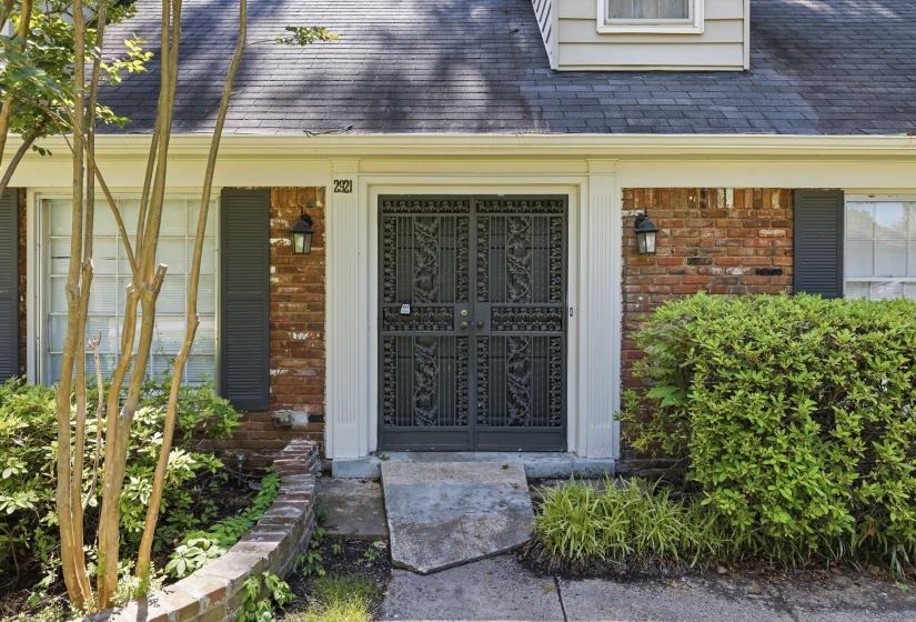 Brick facade featuring a decorative iron entry door, column-flanked entryway, exterior sconce lighting, and dark louvered shutters