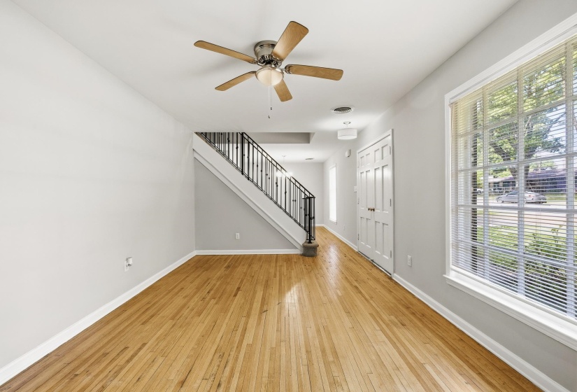 Room featuring wood-finish flooring, a ceiling fan with integrated lighting, a staircase with black iron balusters, white paneled doors, and a large window with horizontal blinds