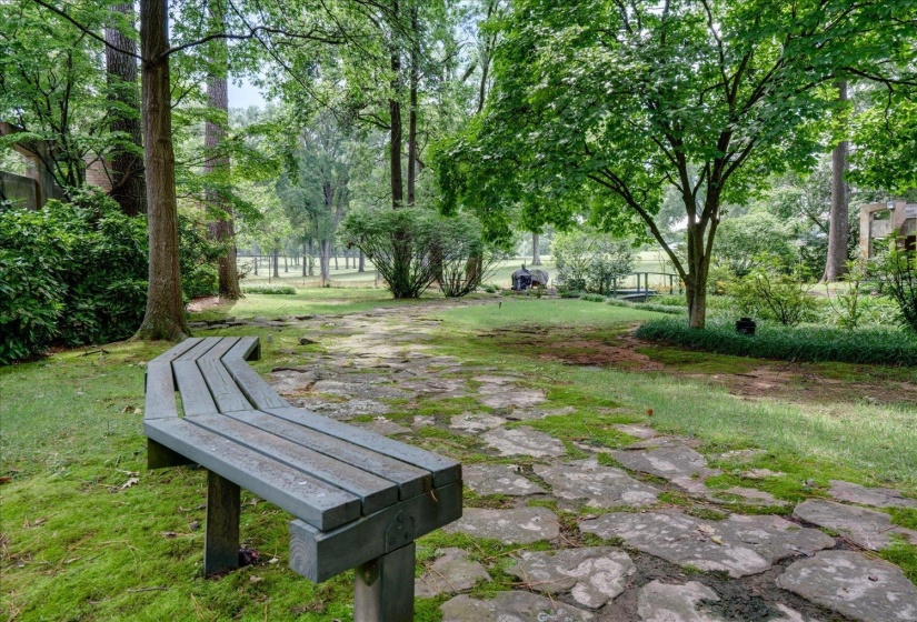 Landscaped grounds featuring a meandering stone pathway, a curved wooden bench, mature trees, and established shrubbery