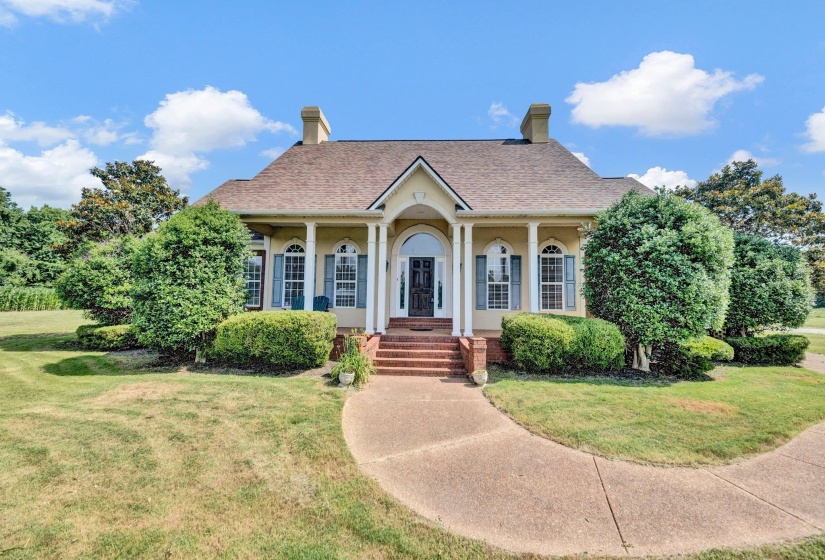 Traditional architecture featuring a covered front porch with white columns, arched windows with shutters, a multi-pane front door, and a brick staircase leading to a concrete pathway