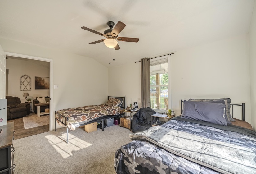 Carpeted interior space featuring a ceiling fan with integrated lighting, natural light from a window, and a doorway opening to a wood-finish floor beyond