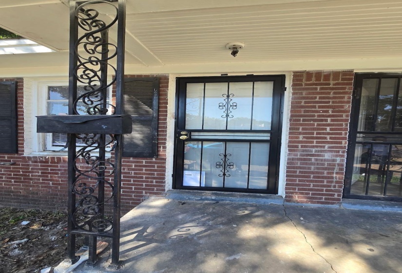 Brick exterior with a covered porch featuring a decorative wrought iron support column, a coordinating wrought iron security screen door, and exterior shutters