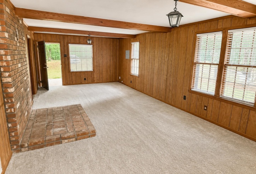 Spacious interior room featuring a brick fireplace, wood-panel walls, exposed ceiling beams, and plush carpet flooring