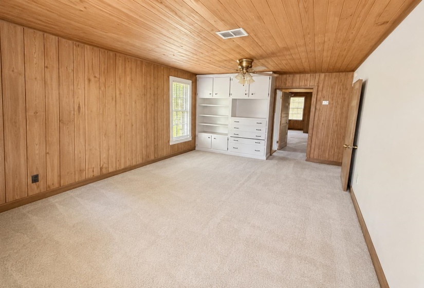 Room featuring wood-finish paneling, light-toned carpet, a window with white trim, built-in white cabinetry with shelving and drawers, and a ceiling fan with integrated lighting