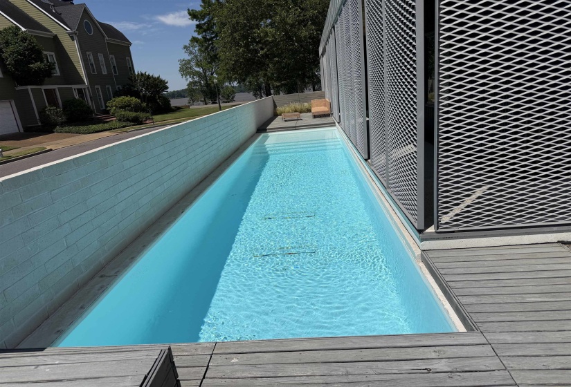 Elongated swimming pool featuring a light blue interior finish, bordered by a textured white wall and a weathered wood-finish deck