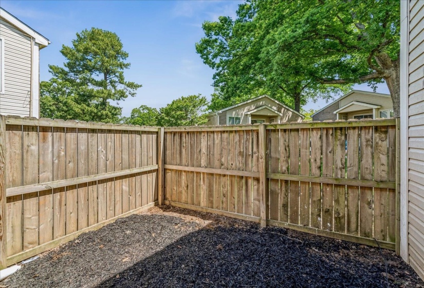 Fenced outdoor area featuring wood panel fencing, dark ground cover, and a partial view of an adjacent structure with siding