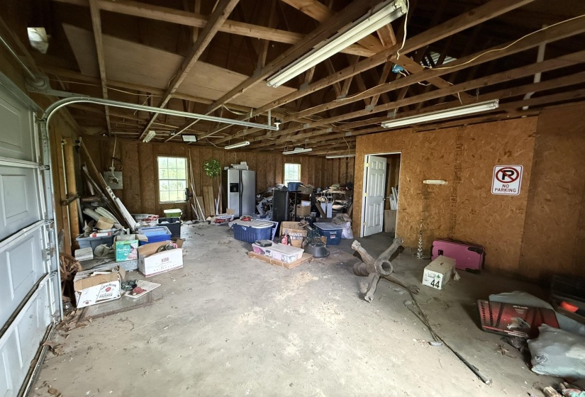 Spacious garage structure featuring exposed wood beam ceiling, concrete flooring, multiple windows, fluorescent lighting, and a white interior door