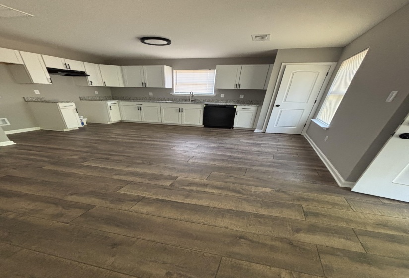 Spacious kitchen featuring wood-finish flooring, white cabinetry with dark hardware, granite-look countertops, a black dishwasher, and a built-in kitchen sink with a window