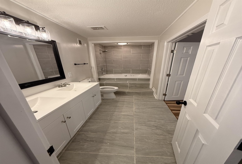 Bathroom featuring a dual-sink vanity with white cabinetry, a built-in jetted tub, a tiled shower surround, and large format floor tiling