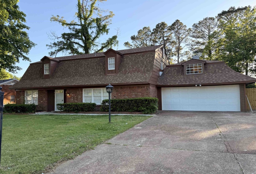 Brick exterior featuring a brown shingle roof with multiple dormer windows, a two-car garage with a white door, a paved driveway, and manicured landscaping