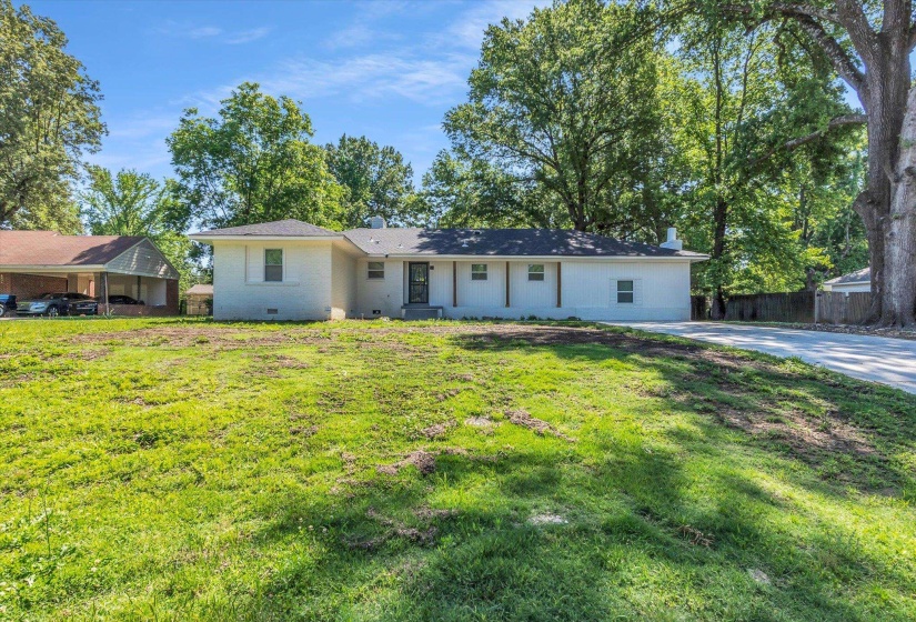 Ranch-style residence featuring a white brick exterior, dark roof, front entry door with glass panel, and mature trees providing natural shade
