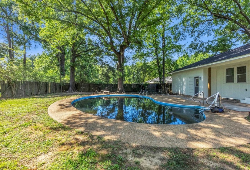 Curved in-ground pool with a surrounding aggregate patio, accompanied by a spacious lawn area and mature trees