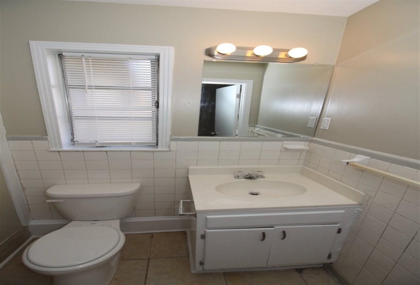 Bathroom featuring white tile walls, a single vanity with a white countertop and base cabinet, a wall-mounted mirror, a three-bulb vanity light, and a window with blinds