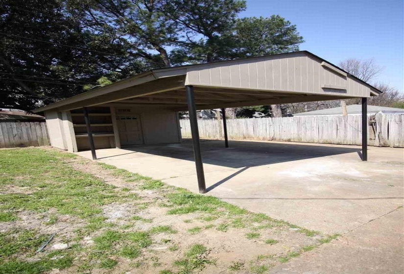 Freestanding carport with a concrete pad and four support posts, featuring an attached storage shed with built-in shelving