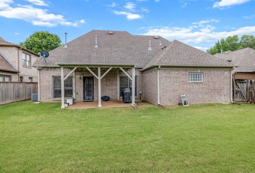 Expansive grassy backyard featuring a covered patio with white support beams, brick exterior, and a shingle roof