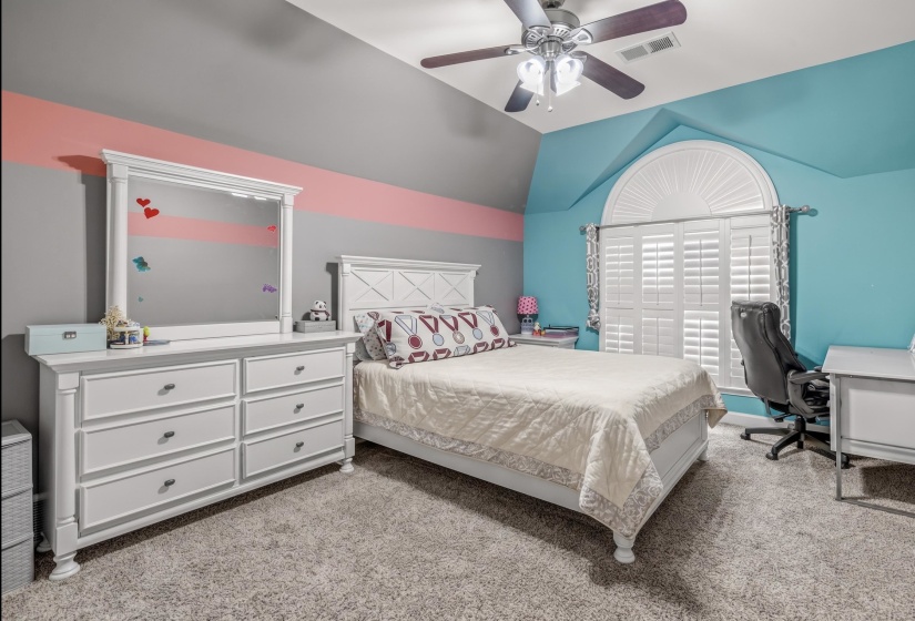 Bedroom featuring a ceiling fan with light fixture, window with arched transom and plantation shutters, and soft carpet flooring
